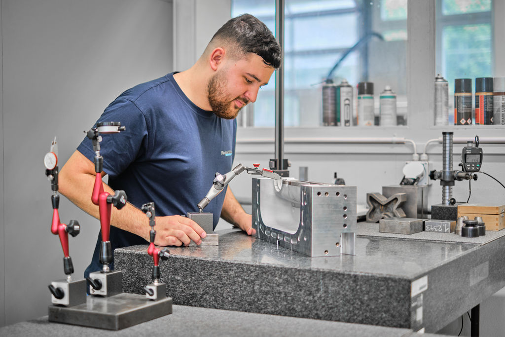 Un homme vêtu d'une chemise bleu marine vérifie un bloc de métal brillant contenant des outils de mesure sur un établi dans un atelier, entouré d'instruments et d'appareils de précision.