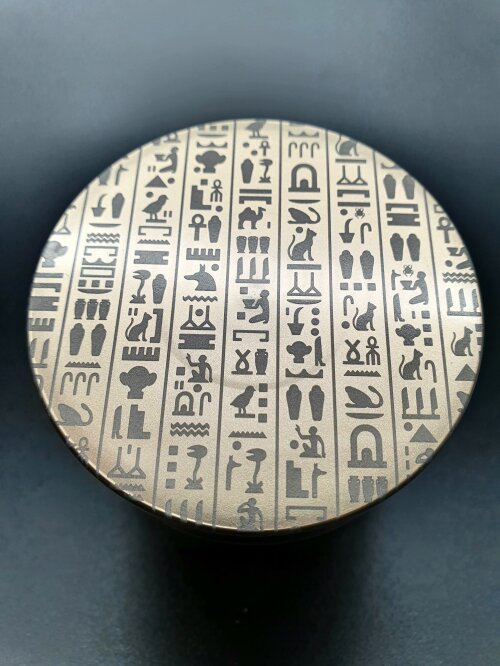 Ein runder Metallgegenstand mit eingravierten altägyptischen Hieroglyphen, die in vertikalen Spalten auf seiner Oberfläche angeordnet sind, vor einem dunklen Hintergrund.