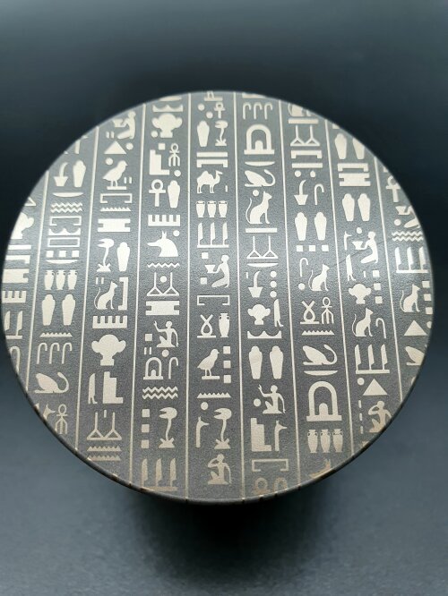 Ein runder Metallgegenstand mit mehreren vertikalen Spalten eingravierter ägyptischer Hieroglyphen vor einem dunklen Hintergrund.