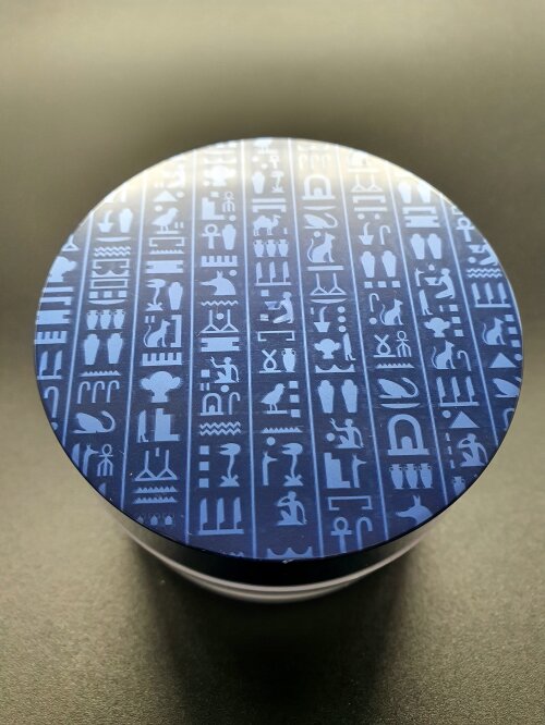 Ein runder, glänzender Behälter mit einem Deckel, der mit blauen und silbernen ägyptischen Hieroglyphen vor einem dunklen Hintergrund verziert ist.
