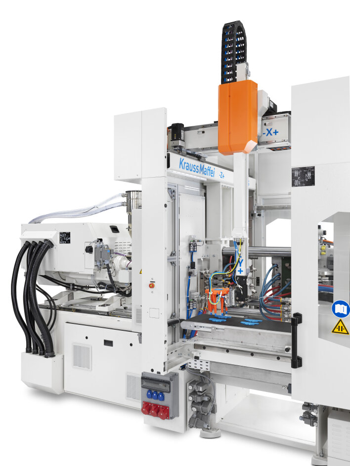 Eine weiße Industriespritzgießmaschine von KraussMaffei mit sichtbaren Kabeln, Schalttafeln und einem orangefarbenen Roboterarm an der Oberseite. Die Maschine ist mit Sicherheitsschildern und blauer Beschriftung ausgestattet.