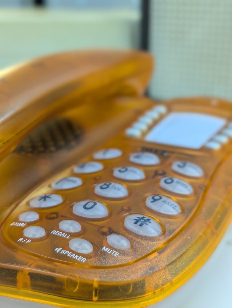 Nahaufnahme eines orangefarbenen, durchsichtigen Festnetztelefons, das die Tastatur mit weißen Tasten und Aufschriften wie MUTE, RECALL und SPEAKER zeigt. Das Telefon ist leicht angewinkelt und der Fokus liegt auf den Tasten.