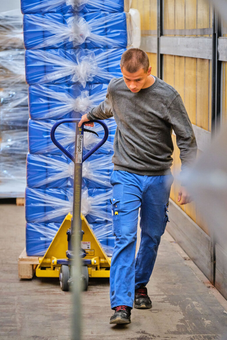 Un homme vêtu d'un pantalon de travail bleu et d'un pull gris utilise un transpalette pour déplacer une grande pile de paquets bleus emballés à l'intérieur d'un entrepôt.