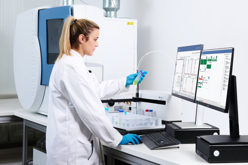 Ein Wissenschaftler, der einen weißen Kittel und blaue Handschuhe trägt, bedient Laborgeräte und analysiert in einem Labor die auf Computerbildschirmen angezeigten Daten.
