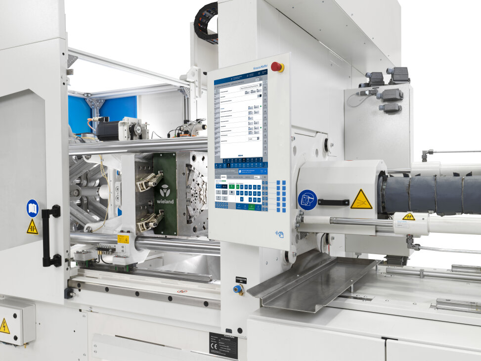 Nahaufnahme einer industriellen Spritzgießmaschine mit offener Form, Bedienfeld mit Bildschirm, Warnschildern und sichtbaren mechanischen Teilen.