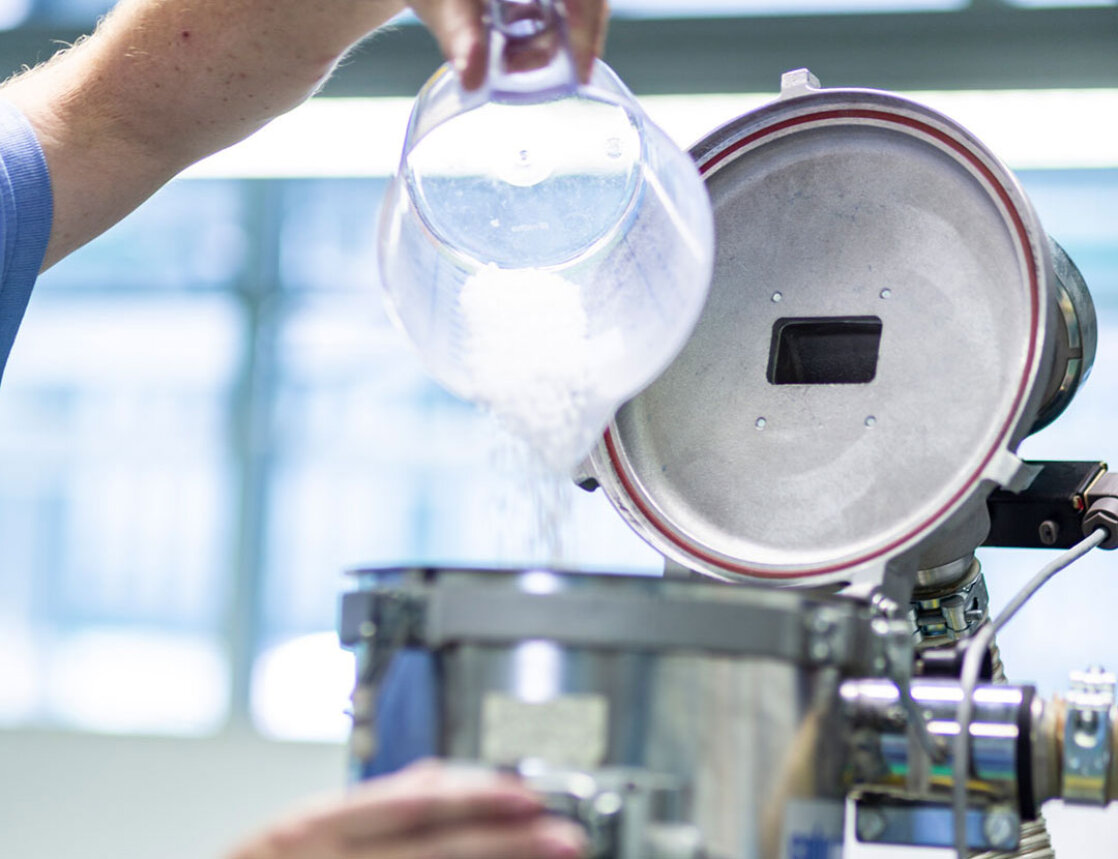 Eine Person schüttet weißes Granulat aus einem durchsichtigen Behälter in eine Industriemaschine aus rostfreiem Stahl mit offenem Deckel, wahrscheinlich Teil eines Herstellungs- oder Laborprozesses.