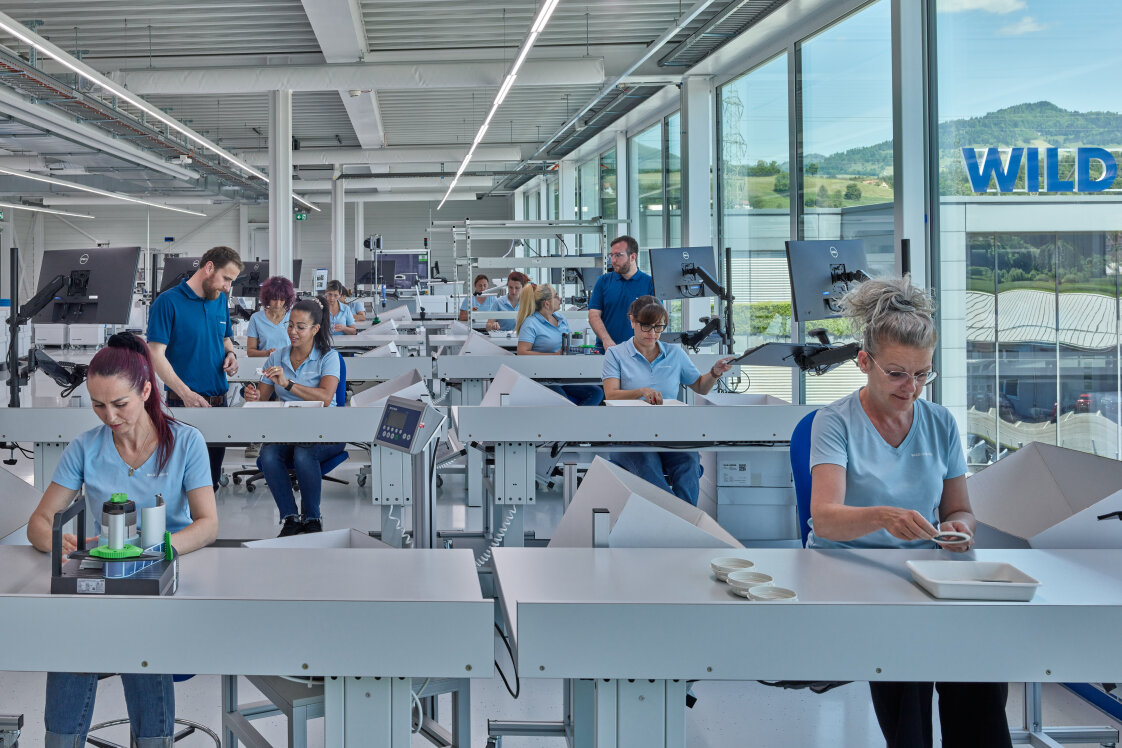 Eine Gruppe von Personen in hellblauen Uniformen arbeitet an einzelnen weißen Schreibtischen in einer hellen, modernen Fabrik oder einem Labor mit großen Fenstern und natürlichem Licht. Draußen sind grüne Hügel zu sehen.