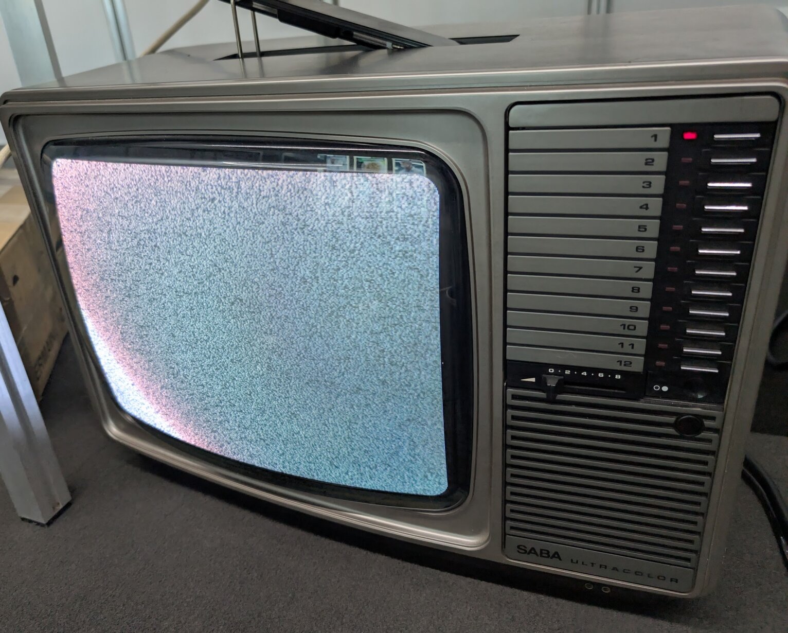 Ein alter Fernseher mit statischem Bild, silbernem Gehäuse, nummerierten Kanaltasten auf der rechten Seite und dem Markennamen SABA ultracolor auf der Unterseite. Der Fernseher steht auf einem Teppichboden.