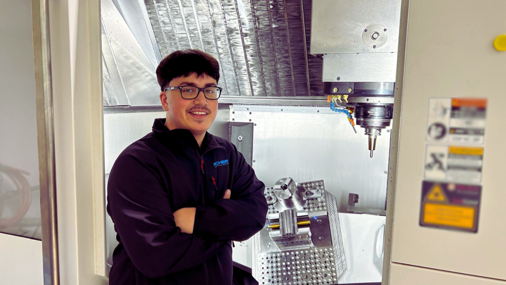 Eine Person mit dunklem Haar und Brille, die eine schwarze Jacke trägt, steht lächelnd mit verschränkten Armen vor einer CNC-Maschine und einem Metallteil in einer Industrieumgebung.