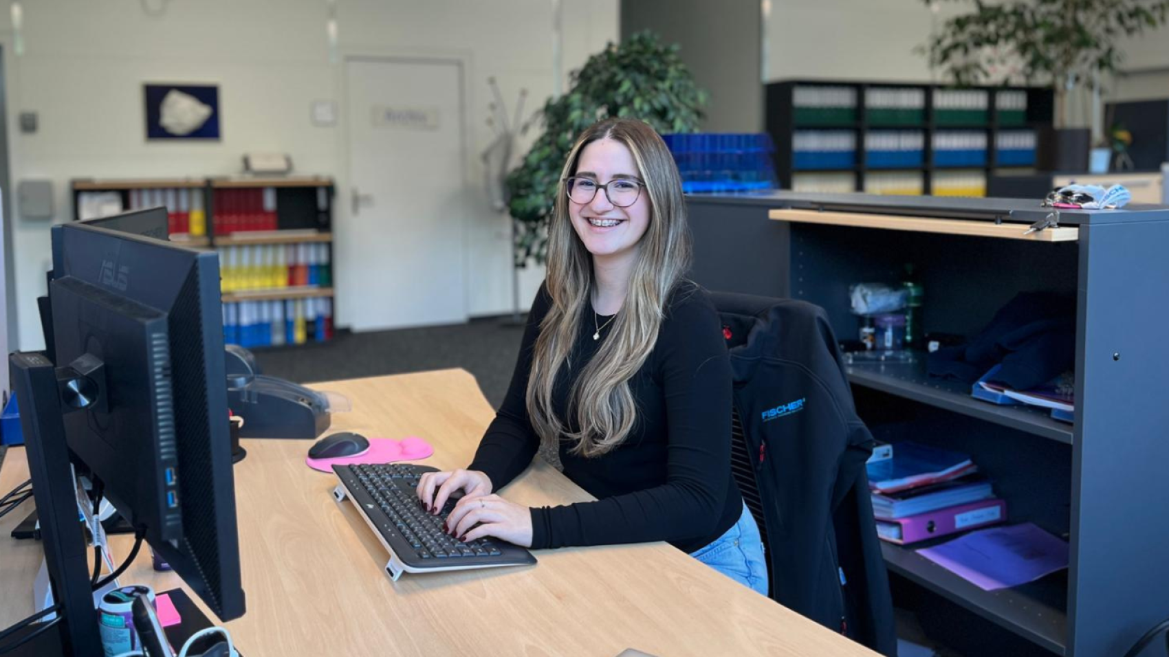 Eine junge Frau mit langen Haaren und Brille lächelt, während sie an einem Schreibtisch in einem Büro sitzt und auf einer Tastatur tippt. Im Hintergrund sind Regale mit bunten Aktenordnern und Pflanzen zu sehen.