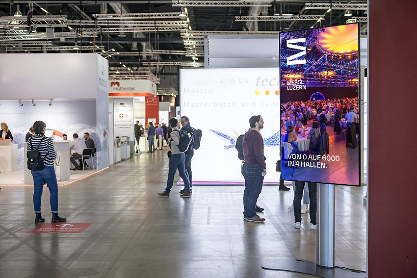 Ein grosser digitaler Bildschirm in einer Messehalle zeigt Werbung für die Messe Luzern mit dem Text 'Von 0 auf 6000 in 4 Hallen'. Im Hintergrund sind Messestände und Besucher sichtbar.