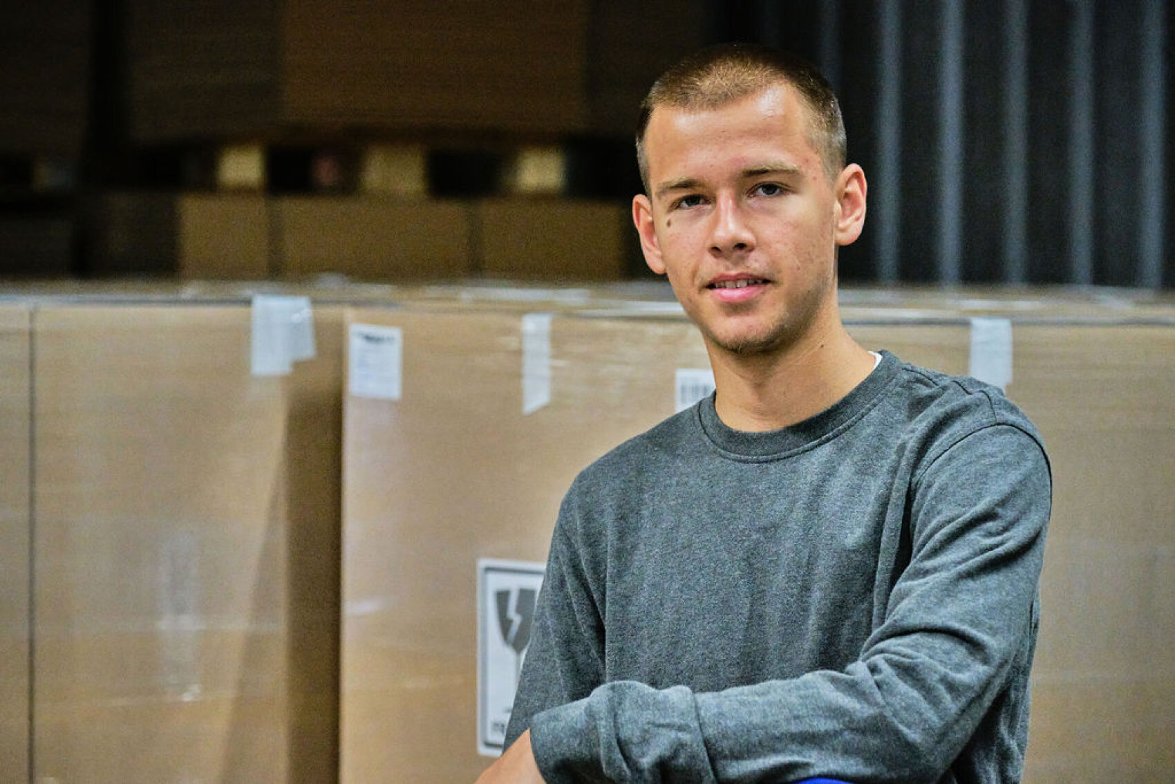Ein junger Mann mit kurzen Haaren und einem grauen langärmeligen Hemd sitzt vor gestapelten Kartons in einer Lagerhalle, blickt in die Kamera und lächelt leicht.