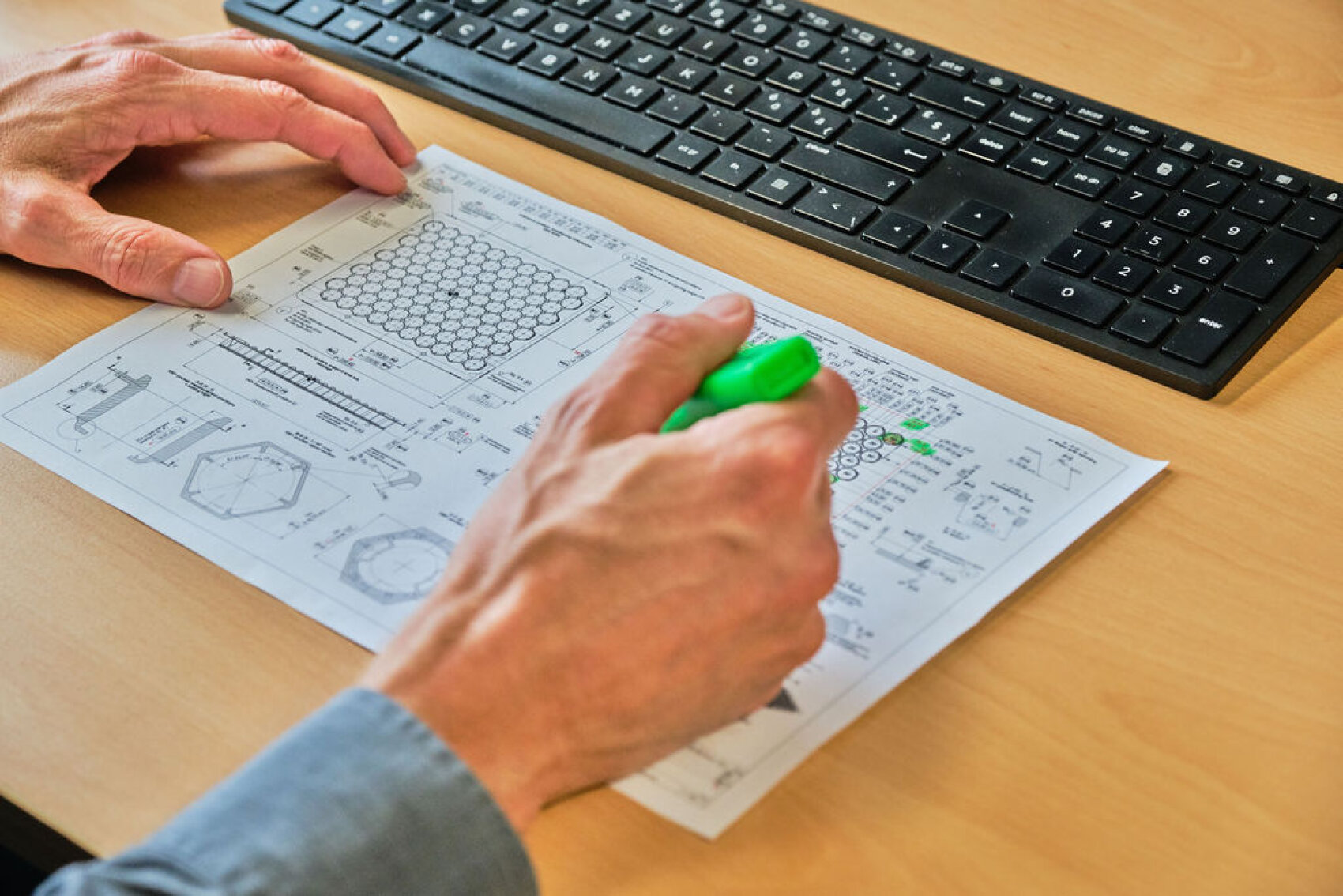 Eine Person markiert Teile einer technischen Zeichnung mit einem grünen Marker an einem Schreibtisch, in dessen Nähe sich eine Computertastatur befindet.