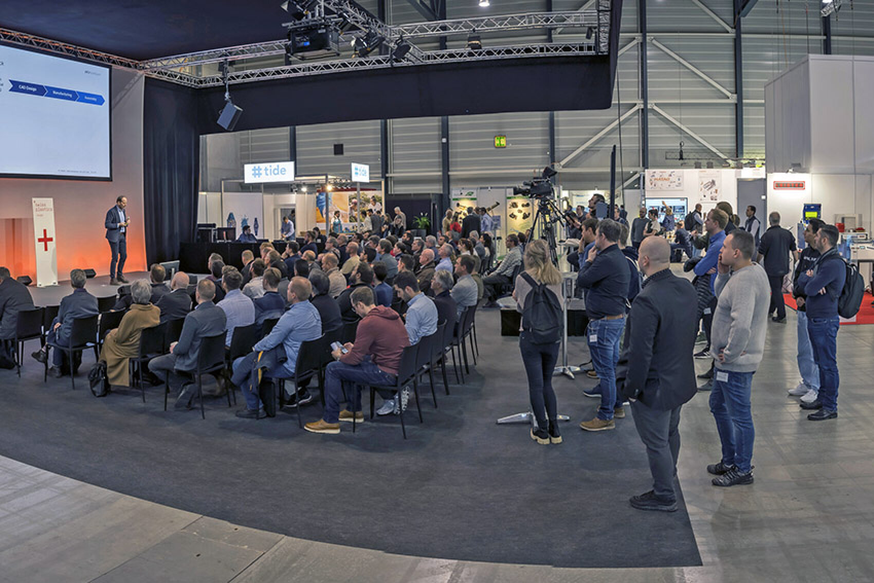 Ein Redner präsentiert auf einer Konferenz vor einem sitzenden Publikum. Mehrere Personen stehen im hinteren und seitlichen Teil des Raumes. Im Hintergrund des Veranstaltungssaals sind Messestände und Banner zu sehen.