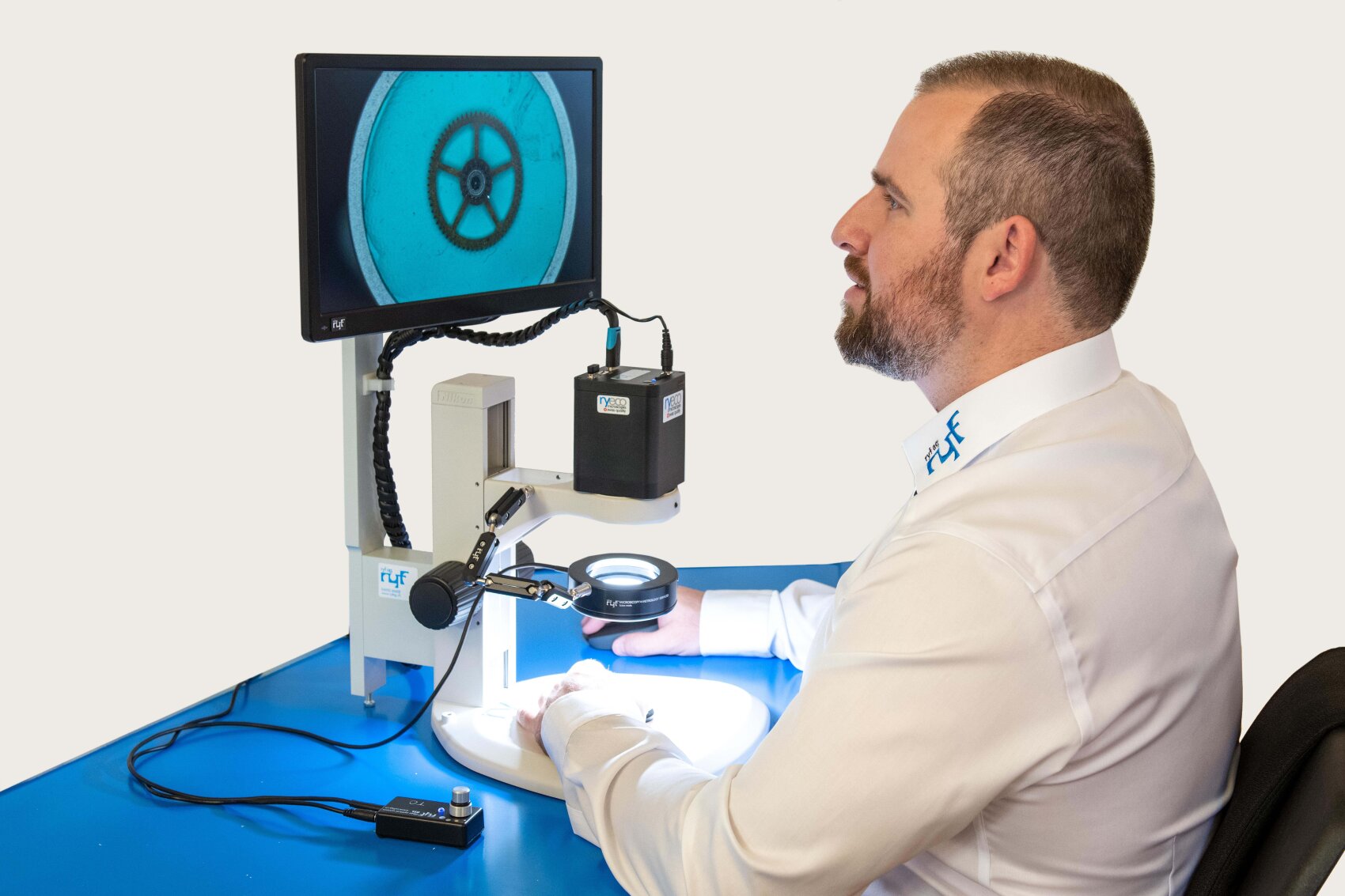 Ein Mann in einem weißen Hemd untersucht mit einem Digitalmikroskop ein rundes Objekt, das auf einem Monitor zu sehen ist, während er an einem blauen Arbeitsplatz sitzt.